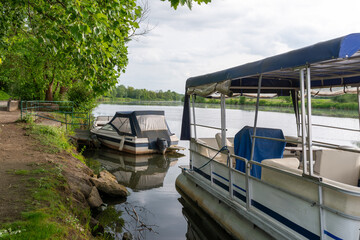 Covered motorboats and a pontoon boat are moored along a quiet riverbank under leafy trees. The calm water and soft light create a tranquil atmosphere associated with relaxation, slow travel, and natu © Volha