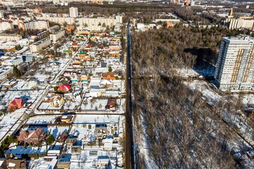 Fototapeta premium Winter drone view of snow-dusted houses flanking a long road, meeting a bare forest. Urban edge contrasting with frosty nature under bright skies