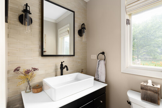 A small half bathroom with a brown subway tile accent wall, white vessel sink on a marble countertop and black cabinet, and black sconces.
