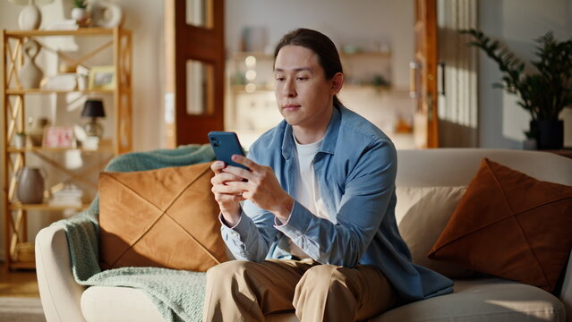 Asian guy typing smartphone looking screen at couch comfortable house closeup