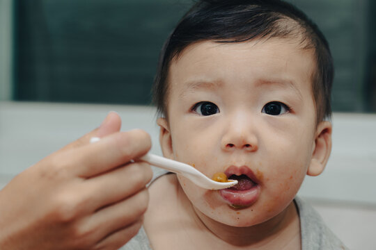 An expressive close-up of a content Asian baby enjoying mealtime, being fed by a spoon. A curious baby eating with a spoon. A joyful Asian infant being spoon-fed, showing a messy face and big eyes.
