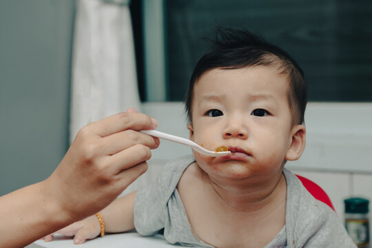 An expressive close-up of a content Asian baby enjoying mealtime, being fed by a spoon. A curious baby eating with a spoon. A joyful Asian infant being spoon-fed, showing a messy face and big eyes.