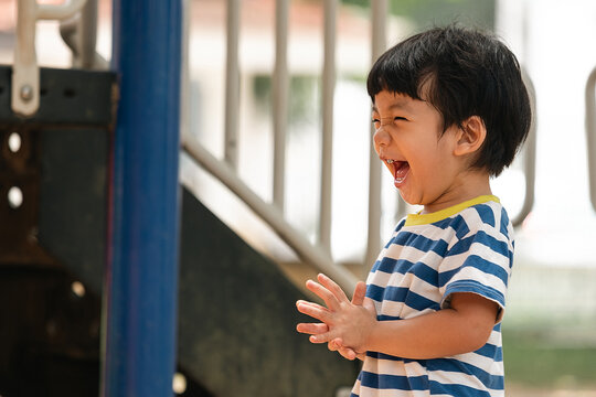 Joyful little Asian boy laughing loudly while playing at the outdoor playground. Happy Asian toddler clapping his hands at a public park playground. Pure childhood joy.