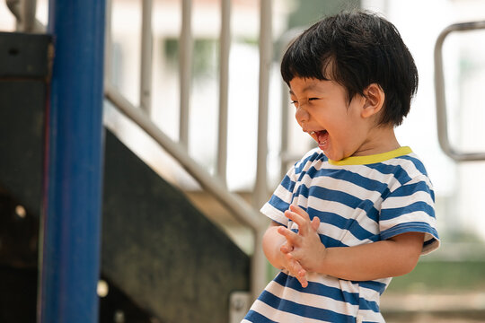 Joyful little Asian boy laughing loudly while playing at the outdoor playground. Happy Asian toddler clapping his hands at a public park playground. Pure childhood joy.