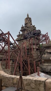 Static shot of a roller coaster cart ascending a steep incline against a backdrop of a stone temple-like structure