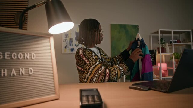 Waist up slowmo of African American shop assistant examining colorful vintage clothing sitting behind wooden counter with laptop in cozy secondhand shop with warm lighting and creative decor