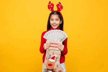 Merry little girl wear red clothes decorative deer horns on head posing hold stocking sock fan of cash money isolated on plain yellow background. Happy New Year Christmas celebration holiday concept.
