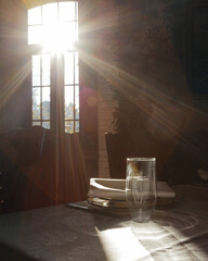 Sunlit rustic dining room with wooden table, plates, glass, and dramatic light streaming through a multi-paned window