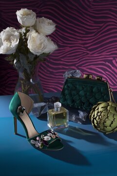 Zapato de tac&oacute;n verde esmeralda con pedrer&iacute;a junto a bolso y perfume, Mockup para campa&ntilde;a de accesorios y calzado de lujo, Bodeg&oacute;n de lujo con zapatos, bolso, perfume y alcachofa sobre fondo azul