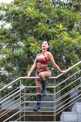 A beautiful, dark-haired Latin American woman dressed as a zombie, wearing a red and black leather top and shorts,black fishnet stockings and black boots.The woman is leaning on the staircase railing.