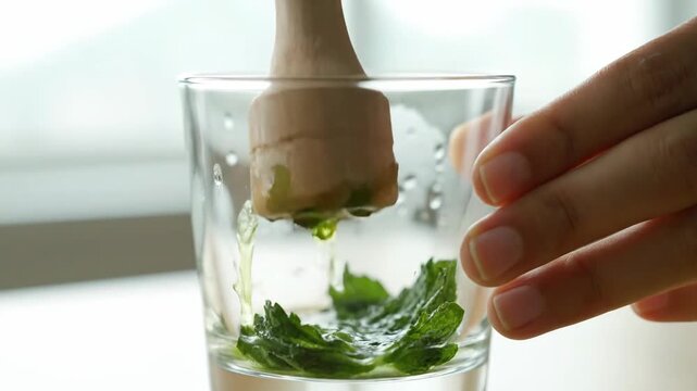 Close-up of person muddling fresh mint leaves in glass, preparing mojito, cocktail, or beverage at home