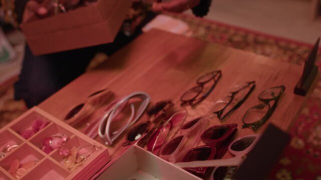 Slowmo tilt up shot of African American male second hand shop assistant organizing vintage eyewear on wooden display table in cozy shop interior with dimmed neon lighting and curated retro accessories