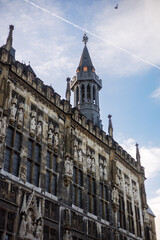 Fototapeta premium Historic gothic town hall facade with stone statues and tall tower against blue sky. European medieval architecture exterior suitable for travel, culture, heritage and city landmark concepts.