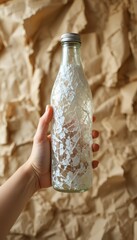 Human hand holds clear glass bottle with white crackle effect design and silver cap Crumpled brown paper background
