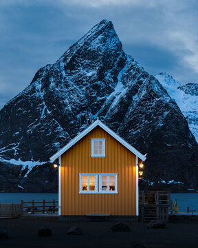 Winter auf den Lofoten in Norwegen mit traditionell gelbem Rorbua in Sakrisoy am verschneiten Fjord und dem Olstinden im Hintergrund zur blauen Stunde unter wolkenverhangenem Himmel bei frostiger Luft