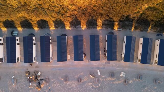 Aerial view of modular beach cabins aligned along a sandy shore, backed by golden coastal vegetation and scattered leisure items, capturing the contrast between geometric order and natural textures.