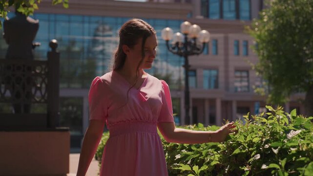 sunlit city garden young woman pink dress wanders along hedge, touches leaves, basks in soft cinematic light frames shift between wide urban plaza and intimate profile closeups moods alternate