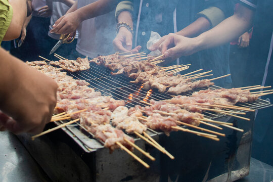 Hands turning savory pork skewers (thit xien nuong) over a smoking charcoal grill at a busy street food stall with students in the background.