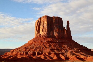 A striking red rock formation rises prominently against a partly cloudy sky in a desert landscape.