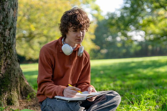 Unhappy person teen sitting under a tree writing in a notebook on sunny day in a large green park