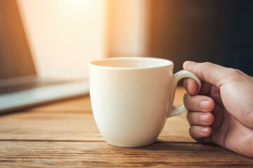 A hand holding a white coffee mug on a wooden table with warm lighting, suggesting a cozy, relaxed atmosphere.