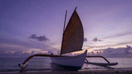 Outrigger canoe at sunset, a carved wooden frame and woven sail silhouetted against a violet tropical sky.