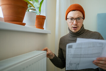 Man with shocked, worried expression holding a utility bill while touching a cold radiator,...