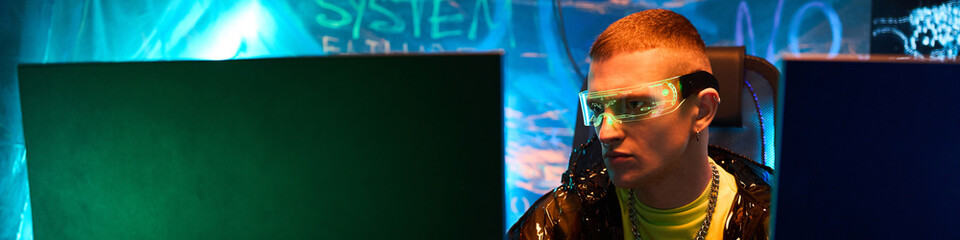 Header of young adult Caucasian man, wearing futuristic glasses, sitting at desk working on computer monitors in dark room, with neon blue lighting and digital graffiti on walls