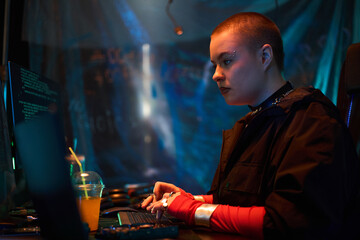 Young adult Caucasian woman with shaved head and facial piercings sitting at desk typing on keyboard, concentrating on computer screens with code in dark room, plastic cup nearby