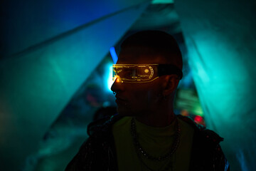 Young adult Caucasian man wearing futuristic illuminated visor standing in dark room with neon blue and green lighting, facing sideways, displaying hacker and cyber club concept
