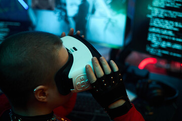 Young adult Caucasian woman wearing virtual reality headset, sitting at computer desk, using VR technology with hacking code on monitor, in dark room with cyber security theme