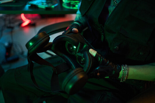 Young male hacker or gamer, wearing black gloves holding virtual reality headset in dimly lit room with computer equipment, showing close up of hands and device, suggesting cybercrime activity - Powered by Adobe