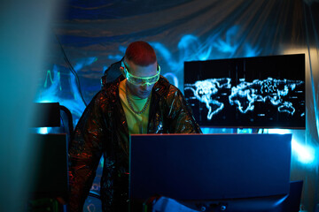 Young adult Caucasian man wearing protective glasses operating multiple computer monitors in dark room, with digital world map on screen, suggesting cybersecurity or hacking activity