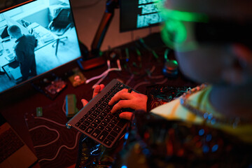 Young adult Caucasian man wearing futuristic glasses sitting at desk typing on wireless keyboard while looking at computer monitor displaying surveillance footage and code
