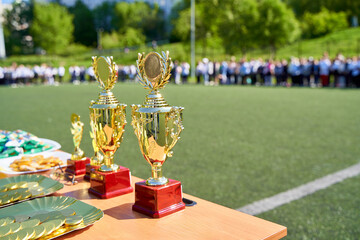 Golden trophies and medals displayed on a table at an outdoor sports event, with participants standing in the background, symbolizing achievement, competition, and celebration.