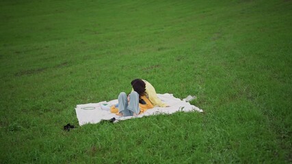 woman alone on blanket in grass, head bowed reading or daydreaming, vast emerald field with gentle breeze, contemplative quiet, mindfulness vibe, cinematic wide shot, pastoral hideaway mood,