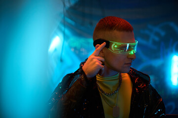 Young adult Caucasian man wearing futuristic neon visor, touching side of smart glasses, standing in cyberpunk environment, with blue and green lighting suggesting hacker activity