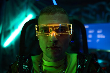 Portrait of young adult Caucasian man wearing futuristic cyberpunk neon visor sitting in dark room with glowing screens, facial piercings visible, looking directly at camera, hacker concept