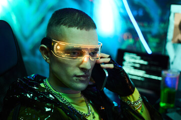 Young adult Caucasian man wearing futuristic glasses sitting at computer desk talking on smartphone surrounded by digital screens with code displaying hacker activity