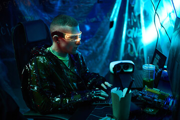 Young adult Caucasian man wearing futuristic glasses, sitting at desk typing on laptop, surrounded by electronic components and VR headset, in dark room with neon lighting