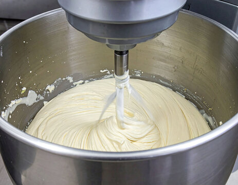 This industrial stainless steel stand mixer whips smooth, creamy yellow batter in a professional kitchen setting. Ideal for bakery marketing, culinary blogs, and food production concepts.