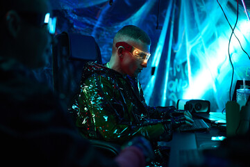 Obraz premium Young adult Caucasian man wearing futuristic glasses sitting at computer desk typing on keyboard in dark room with neon blue lighting, another partially visible in foreground