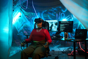 Young adult Caucasian woman wearing virtual reality headset sitting in dark room surrounded by computer monitors displaying code and hacking tools, wires and neon graffiti visible