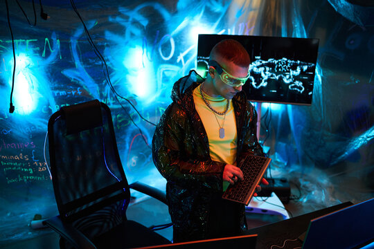 Young adult Caucasian man, wearing futuristic glasses, standing in dark room with neon lights, holding wireless keyboard and looking at computer monitor displaying digital world map - Powered by Adobe