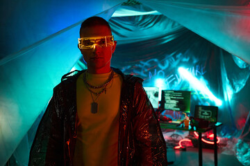 Young adult Black man wearing futuristic glasses standing in front of computer monitors displaying code in dark room with neon lighting, embodying hacker concept, not looking at camera
