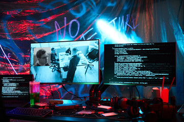 Multiple computer monitors displaying surveillance footage and lines of code on cluttered desk, showing hacker workspace with visible keyboard, headphones, and illuminated background