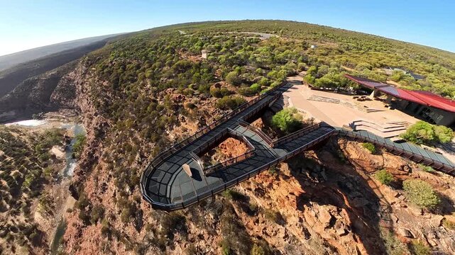 The Kalbarri Skywalk walkway over the Murchison River gorge, offering a panoramic view of rugged red rocks, sparse shrubs, and a vivid blue sky. Ideal for travel, adventure, and nature imagery.