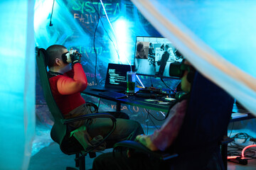 Two young adult men sitting at desks wearing virtual reality headsets and using computers in dark room with neon blue lighting, digital code and security warnings projected on walls