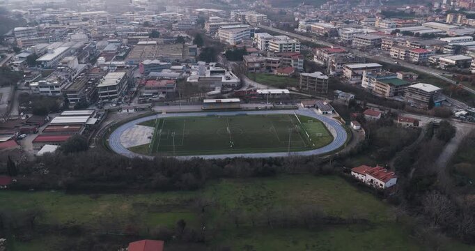 Aerial view of a stadium with players, surrounded by tanning factory buildings and roads, presenting a blend of urban and recreational spaces, Solofra, Campania, Italy.