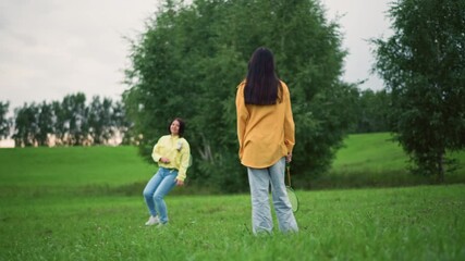 Two women playing badminton in green meadow under overcast sky, casual yellow jackets and denim jeans, spirited rally with racket and shuttlecock, trees lining horizon, lush grass, relaxed weekend